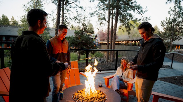 Four friends enjoy a campfire outdoors at dusk, sitting and standing around, with smiles and laughter.