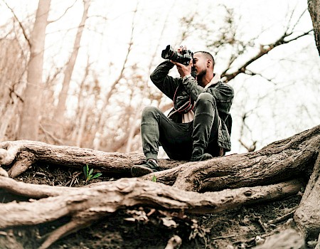A person is sitting on tree roots in a forest, taking a photo with a DSLR camera.