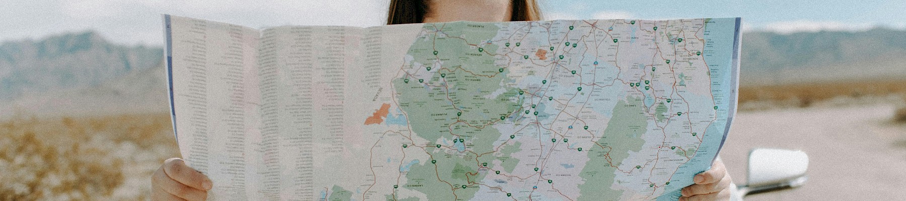 A woman stands in front of a car, holding a map partially covering her face, in a desert landscape under a blue sky.