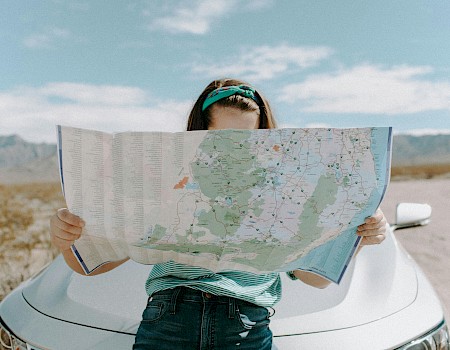 A woman stands in front of a car, holding a map partially covering her face, in a desert landscape under a blue sky.