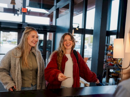 Two young women are smiling and interacting with a front desk agent at the reception desk of Riverhouse Lodge in Bend, OR in a brightly lit, modern store or cafe.