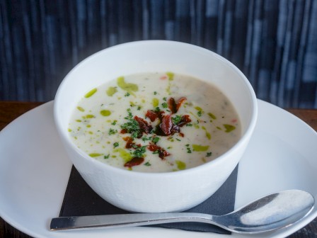 A bowl of creamy soup garnished with herbs, chili, and a drizzle of oil, served on a white plate with a spoon on a wooden table. Daily soups created at Currents at Riverhouse Lodge in Bend, Oregon
