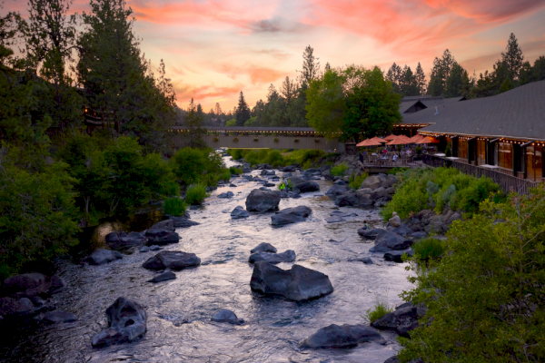 A peaceful river scene at sunset with trees, rocks, a bridge, and a cozy outdoor restaurant along the bank.
