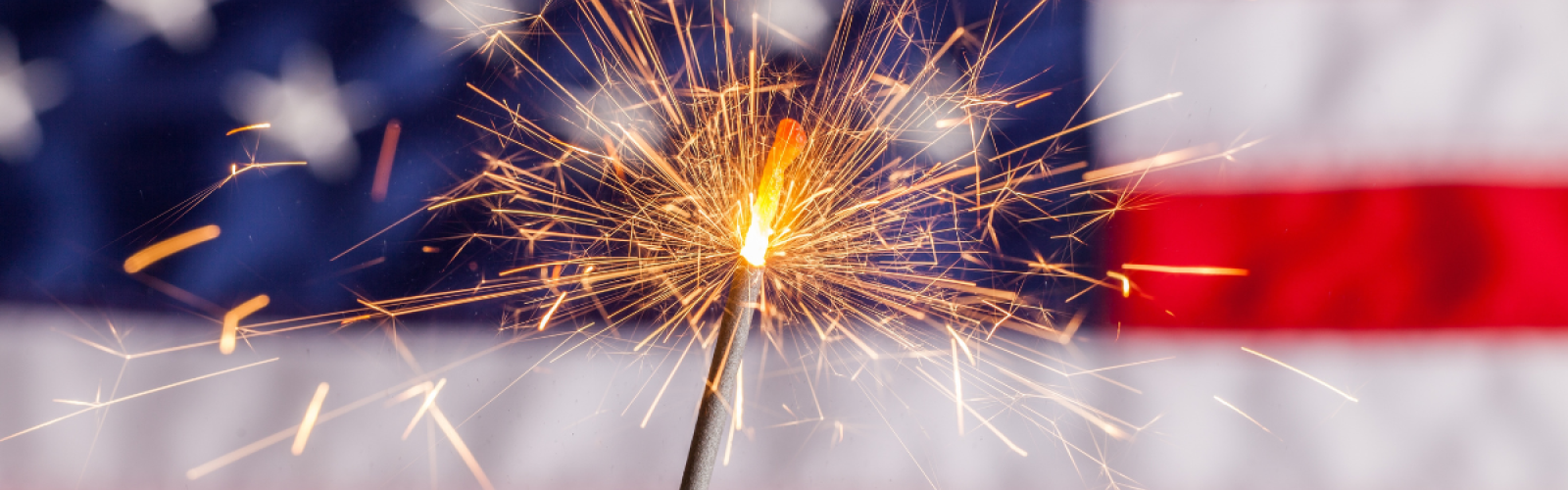 A burning sparkler in focus with an American flag in the blurred background.