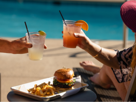 two-people-eating-a-burger-and-clinking-cups-at-riverhouse-lodge-pool-for-fourth-of-july-bbq-at-pool