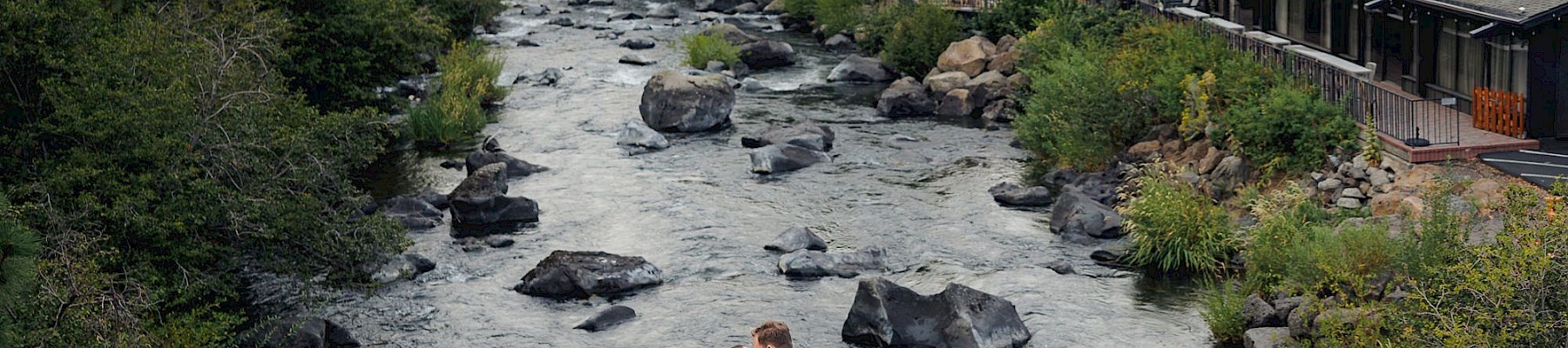 A couple in wedding attire embraces on a bridge over a river, surrounded by trees and a nearby restaurant with outdoor seating.