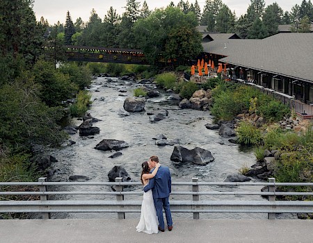 A couple in wedding attire embraces on a bridge over a river, surrounded by trees and a nearby restaurant with outdoor seating.