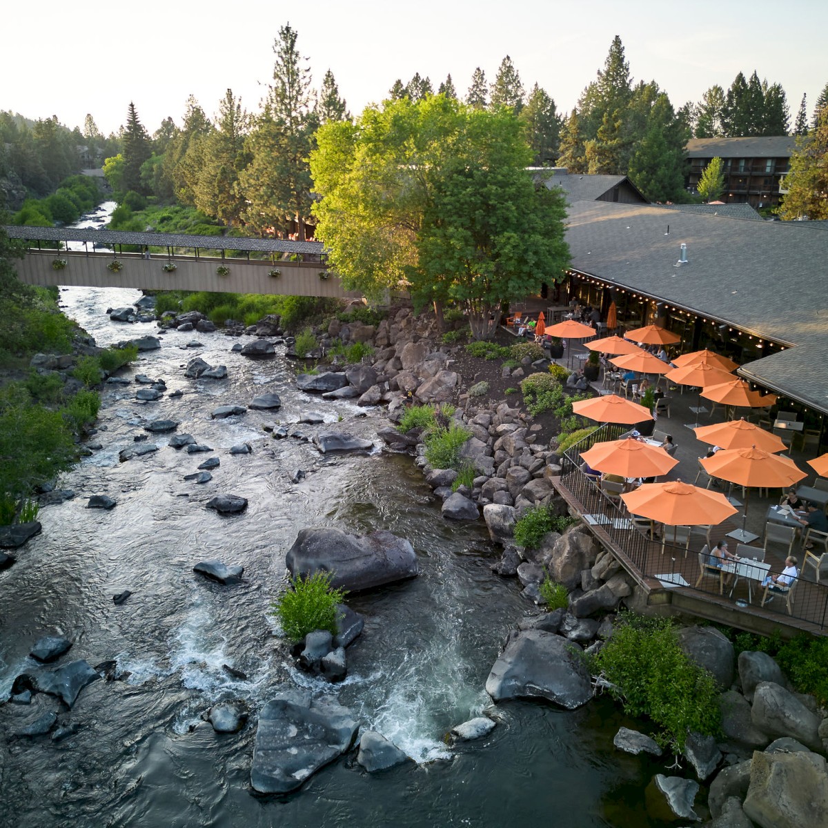 An outdoor restaurant with orange umbrellas beside a flowing river, surrounded by trees and residential buildings in a scenic, green setting.