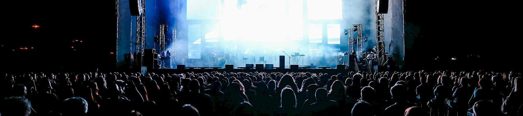 A large crowd watches a brightly lit concert stage at night with speakers and lighting equipment.