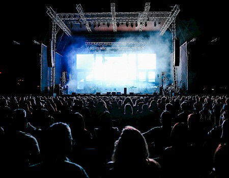 A large crowd watches a brightly lit concert stage at night with speakers and lighting equipment.