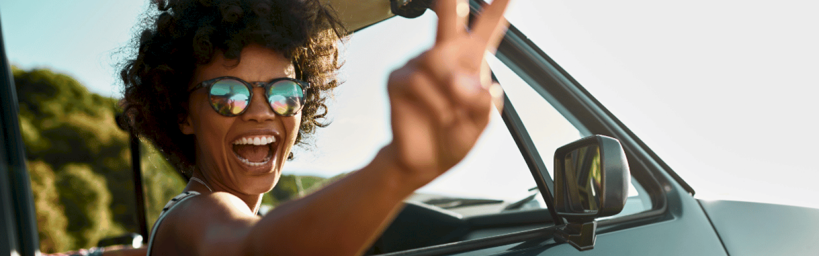 A joyful woman with curly hair and sunglasses waves from a car window, enjoying a sunny outdoor day.