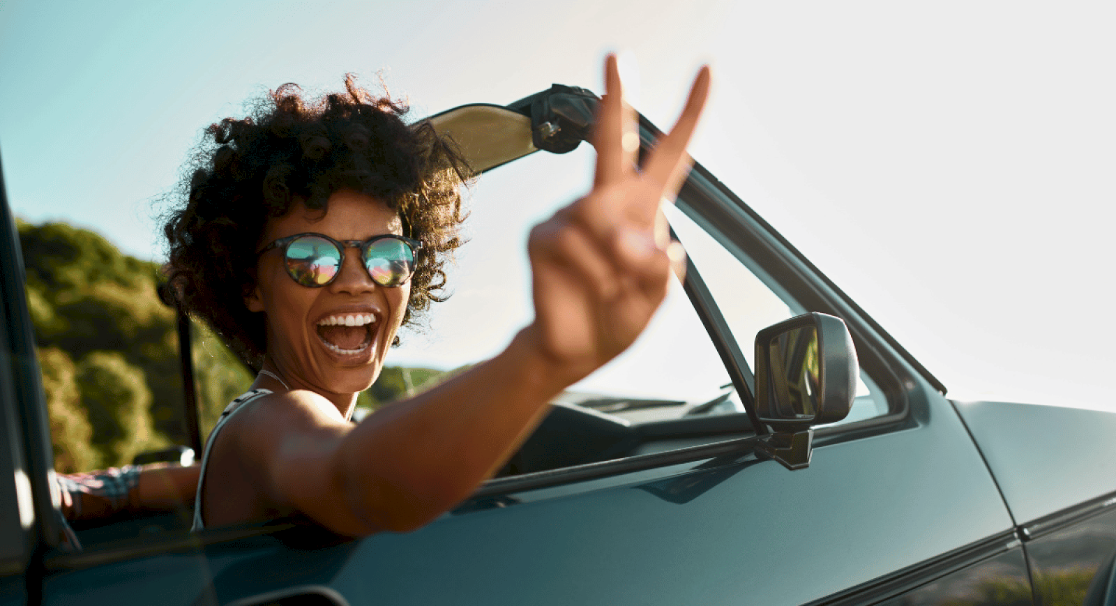 A joyful woman with curly hair and sunglasses waves from a car window, enjoying a sunny outdoor day.
