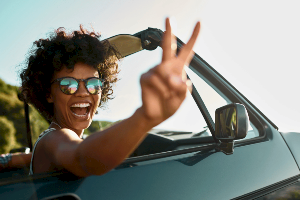 A woman with curly hair and sunglasses is smiling and making a peace sign out of a car window during a sunny day.