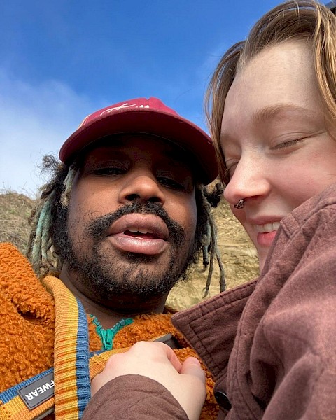 Two people taking a close-up selfie outdoors with blue sky and clouds in the background.