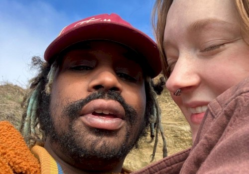 Two people taking a close-up selfie outdoors with blue sky and clouds in the background.