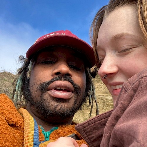 Two people taking a close-up selfie outdoors with blue sky and clouds in the background.
