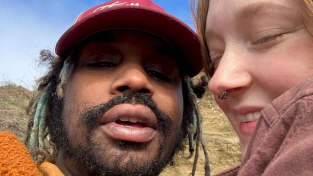 Two people taking a close-up selfie outdoors with blue sky and clouds in the background.