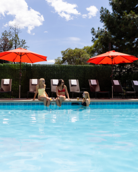Three women sit by a swimming pool with red umbrellas, chatting and relaxing on poolside chairs on a sunny day.