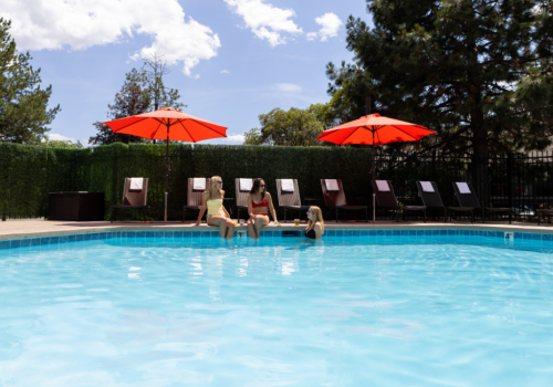 Three women sit by a swimming pool with red umbrellas, chatting and relaxing on poolside chairs on a sunny day.