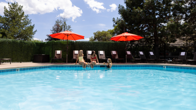 Three women sit by a swimming pool with red umbrellas, chatting and relaxing on poolside chairs on a sunny day.