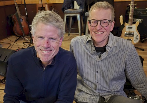 Two male musicians seated indoors with guitars and bass performing acoustic live music at a restaurant stage in Bend Oregon