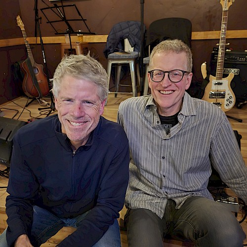 Two male musicians seated indoors with guitars and bass performing acoustic live music at a restaurant stage in Bend Oregon