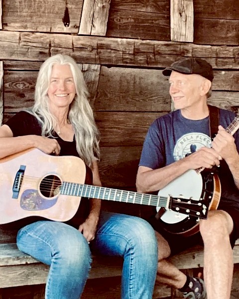 Female guitarist and male banjo player performing acoustic bluegrass music seated outdoors in rustic setting play Currents at Riverhouse on June 12