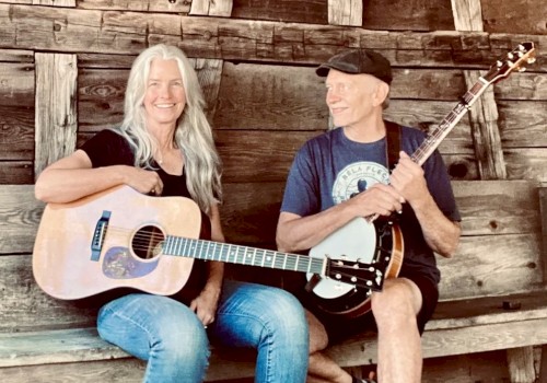 Female guitarist and male banjo player performing acoustic bluegrass music seated outdoors in rustic setting play Currents at Riverhouse on June 12