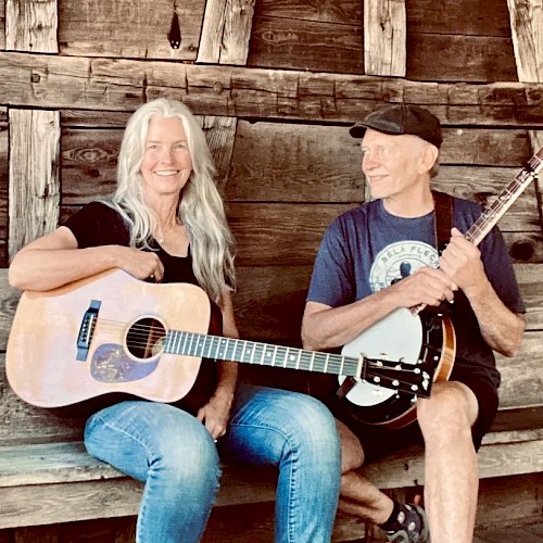 Female guitarist and male banjo player performing acoustic bluegrass music seated outdoors in rustic setting play Currents at Riverhouse on June 12
