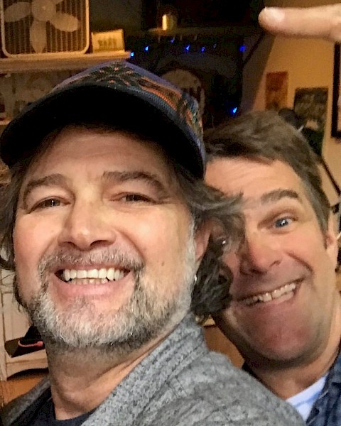 Two male musicians seated indoors smiling with guitars and stage equipment behind them during live brunch performance