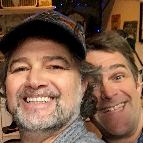 Two male musicians seated indoors smiling with guitars and stage equipment behind them during live brunch performance
