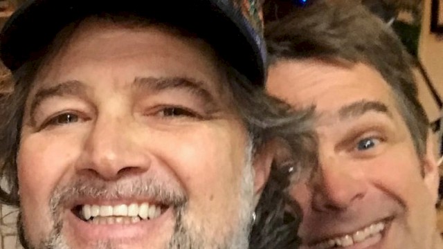 Two male musicians seated indoors smiling with guitars and stage equipment behind them during live brunch performance