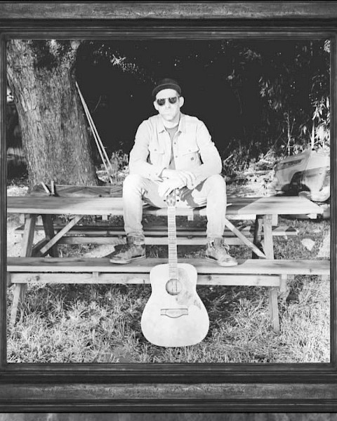 Male musician seated outdoors with acoustic guitar in black and white portrait style setting