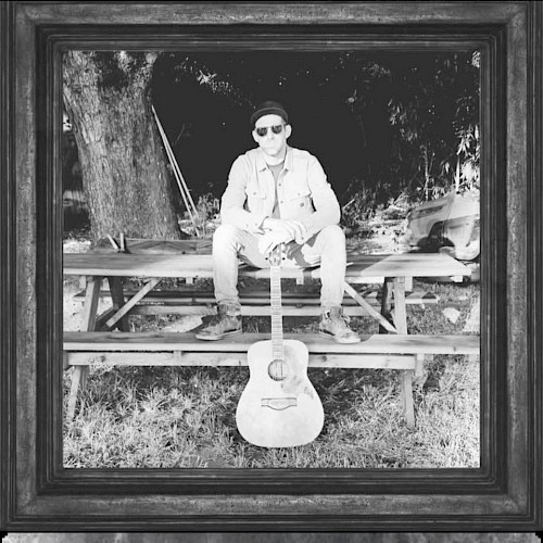 Male musician seated outdoors with acoustic guitar in black and white portrait style setting