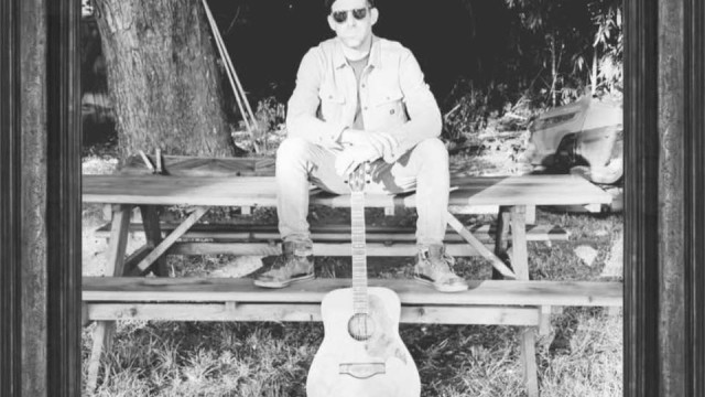 Male musician seated outdoors with acoustic guitar in black and white portrait style setting