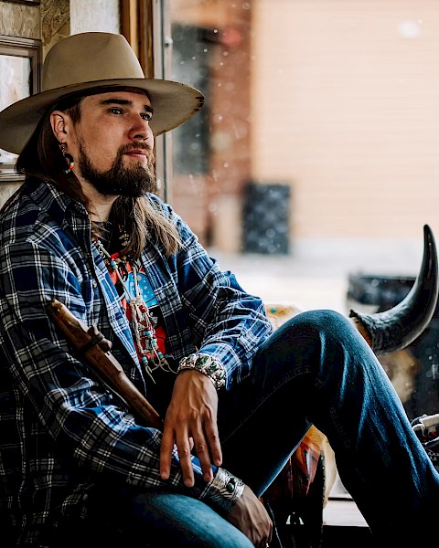 Male musician with long hair and hat seated by window holding instrument wearing patterned jacket during live performance at Currents at Riverhouse in Bend Oregon June 21