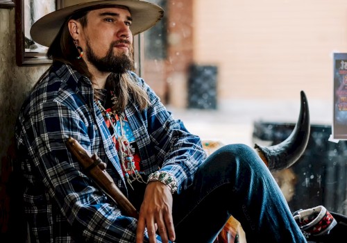 Male musician with long hair and hat seated by window holding instrument wearing patterned jacket during live performance at Currents at Riverhouse in Bend Oregon June 21