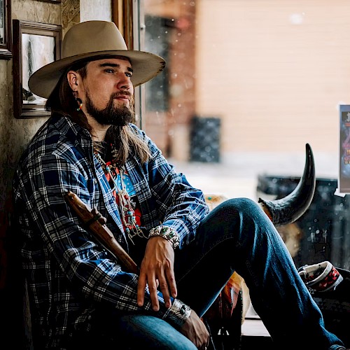 Male musician with long hair and hat seated by window holding instrument wearing patterned jacket during live performance at Currents at Riverhouse in Bend Oregon June 21