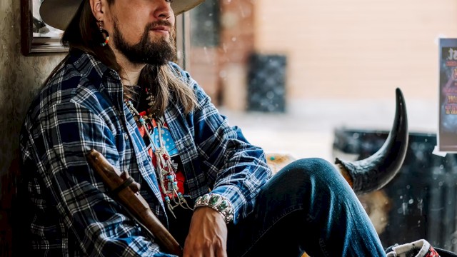Male musician with long hair and hat seated by window holding instrument wearing patterned jacket during live performance at Currents at Riverhouse in Bend Oregon June 21