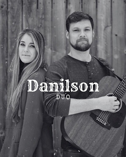 Male and female musicians standing back to back with acoustic guitar in front of wooden wall performing live music at Riverhouse Lodge in Bend Oregon