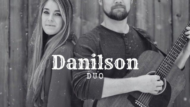 Male and female musicians standing back to back with acoustic guitar in front of wooden wall performing live music at Riverhouse Lodge in Bend Oregon