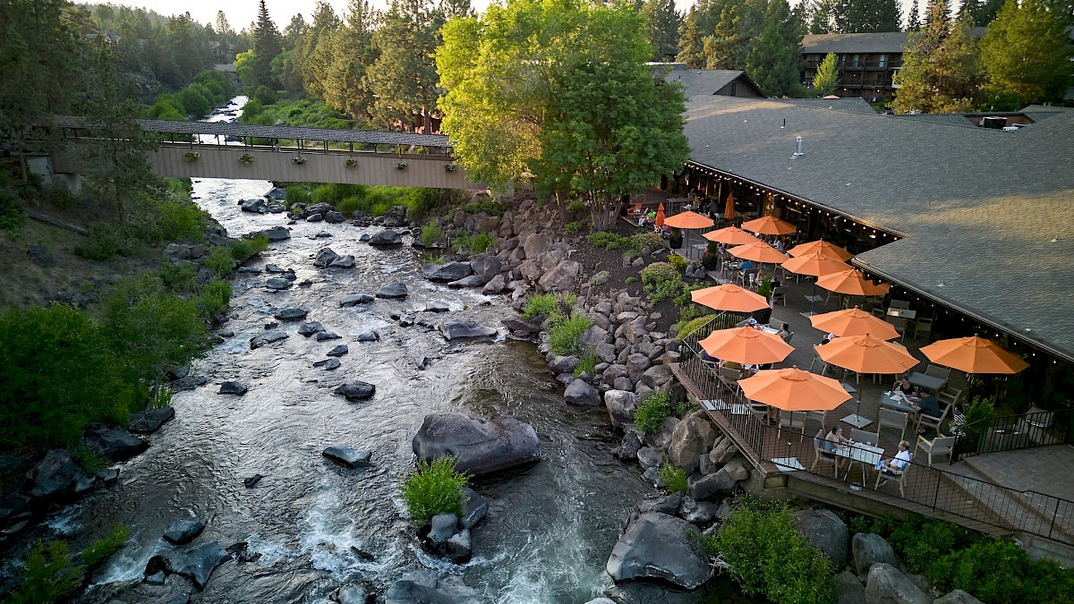 A riverside restaurant with outdoor seating and orange umbrellas, surrounded by rocks, trees, and a bridge over a flowing river.