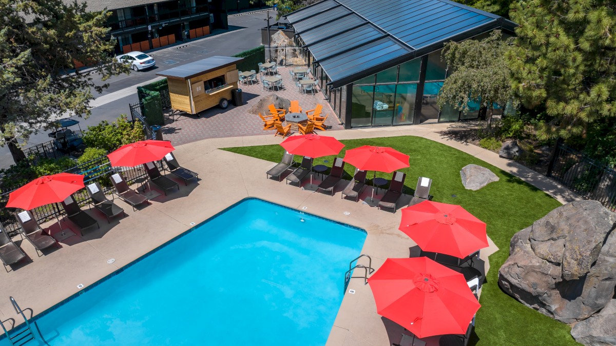 An outdoor swimming pool area with lounge chairs, red umbrellas, a shaded seating area, and a building surrounded by trees.