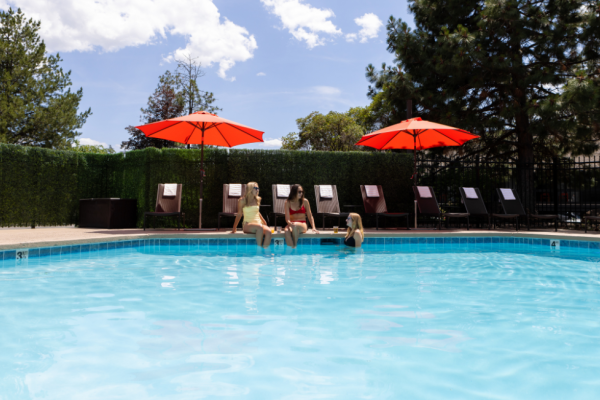 Three women sit by a pool under red umbrellas, enjoying drinks on a sunny day with lounge chairs and trees in the background.