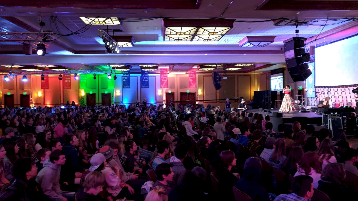 A large audience watches a woman in a colorful dress perform on stage at an indoor event with vibrant lighting.