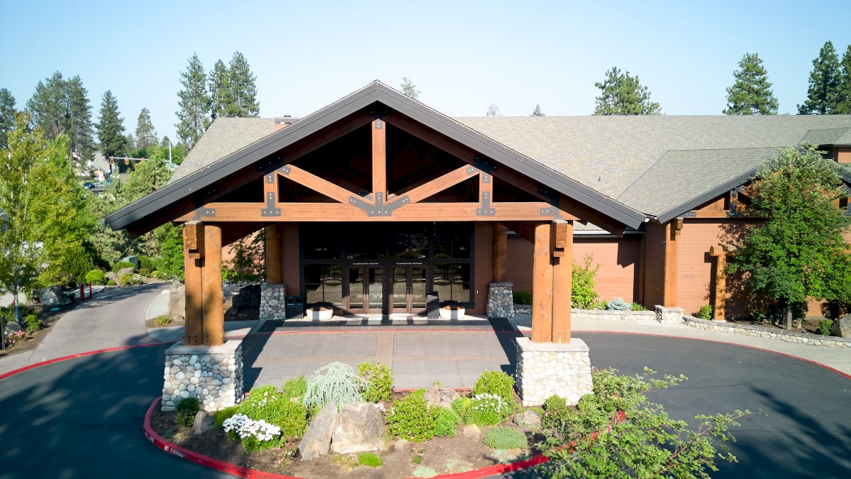 The image shows a wooden lodge or resort building with a large entrance, surrounded by trees and landscaping under a bright blue sky.