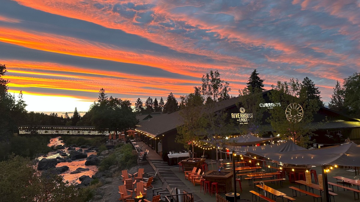 A scenic view of a riverside restaurant at sunset with colorful sky, outdoor seating, string lights, and a bridge over the river.