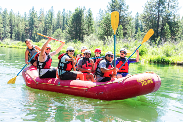 A group of eight people, wearing life jackets and helmets, enjoying a rafting adventure on a calm river surrounded by trees.