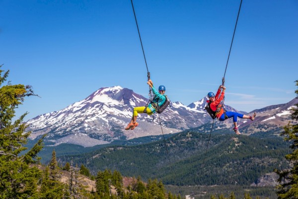 Two people zip-lining in front of a snow-capped mountain under a clear blue sky.