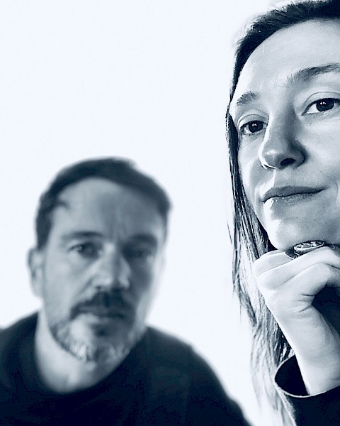 Two people pose for a close-up black and white photo, with one woman in focus and a man blurred in the background.